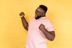 Portrait of positive satisfied man wearing pink shirt raised clenched fists, being happy of winning, expressing triumph, screaming happily. Indoor studio shot isolated on yellow background.