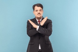 Portrait of angry upset man with mustache standing standing with crossed arms, saying no way, warning, wearing black suit with red tie. Indoor studio shot isolated on light blue background.