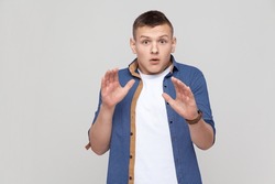 Portrait of scared attractive handsome teenager boy wearing blue shirt gesturing stop with palms and looking surprised with frightened eyes. Indoor studio shot isolated on gray background.