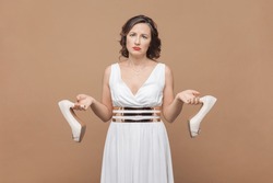 Portrait of sad upset middle aged woman with wavy hair holding elegant shoes on high heels, feels pain on feet, wearing white dress. Indoor studio shot isolated on light brown background.