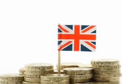 A miniature Union Jack, the flag of Great Britain, stands behind stacks of British Pound Coins on a white background with copy space
