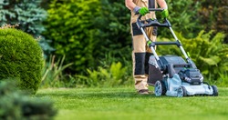 Professional Caucasian Gardener in His 40s Trimming Grass Lawn Using Modern Electric Cordless Mower. Landscaping Industry Theme.