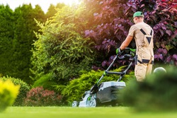 Caucasian Residential Garden Worker in His 40s Trimming Backyard Lawn Using Electric Cordless Grass Mower. Landscaping and Gardening Industry Theme.