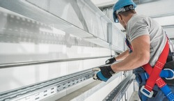Professional Industry Worker Wearing Safety Harness Installing Aluminium Runner Inside Newly Built Commercial Warehouse Building.