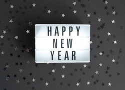Party Time Layout with White Light Box with Black Happy New Year, Black and Silver Confetti of Star Shape on a Black Background. Top-Down View. Flat Lay ideal for Banner, New Years Wishes, Card.
