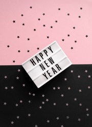 Party Time Layout with White Light Box with Black Happy New Year, Black and Silver Confetti of Star Shape on a Pink-Black Background. Top-Down View. Flat Lay ideal for Banner, New Years Wishes, Card.