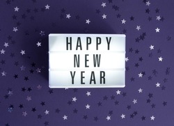Party Time Layout with White Light Box with Black Happy New Year, Black and Silver Confetti of Star Shape on a Dark Violet Background. Top-Down View. Flat Lay ideal for Banner, New Years Wishes, Card.