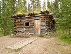 Old Yukon log cabin hidden in the boreal forest (taiga) of Yukon Territory, Canada with chair and table on outside 