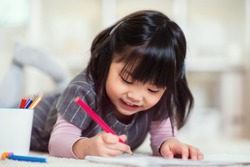 Pretty happy little japanese girl lying and drawing with pencils on white capet at home