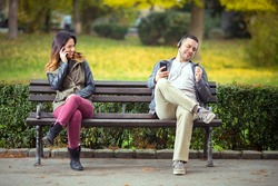 Young man listening to music on his smart phone and singing while woman sitting on the same park bench trying to talk on her phone 