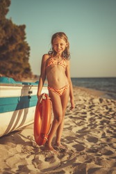 Cute little girl is relaxing on the beach. She is standing next to boat and sunbathing.
