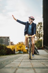 Successful middle-aged businessman waving to someone while riding bicycle on his way to work through the city.
