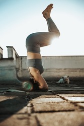 Relaxed woman practicing yoga headstand on a rooftop terrace at sunset.