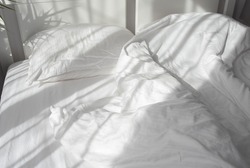Morning light from the window falling on a bed with white satin bedding. 