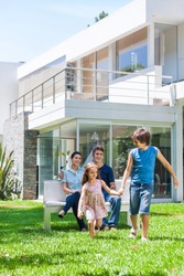 happy family in front of big modern new house outdoors, parents sitting on bench, children running on lawn grass smile