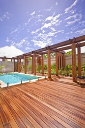 Modern swimming pool with wooden floor and pillars with beam om the poolside patio and garden area under the blue sky with white clouds