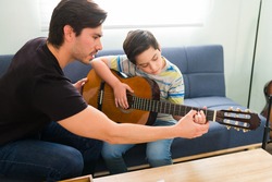 Taking guitar classes with a private music teacher. Hispanic young man helping an elementary boy to play the guitar during home lessons