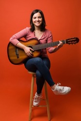 Gorgeous caucasian woman sitting on a stool and learning to play the acoustic guitar against an orange background