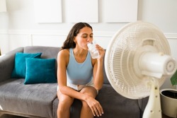 Happy woman trying to cool down drinking an ice cold drink and turning on the electric fan at home