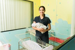 Beautiful happy nurse smiling while carrying a baby in the nursery room of the hospital 