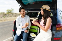 Attractive young man and woman eating a sandwich while resting on the car trunk during a weekend trip together