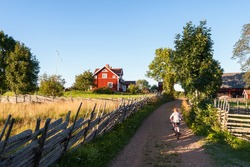 Child riding a bicycle along a small farm lane in rural Sweden pedaling towards a traditional red painted timber house