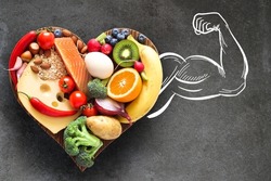 A healthy, balanced diet. Healthy food on a heart-shaped wooden cutting board and a muscular arm as a synonym for strength and health.