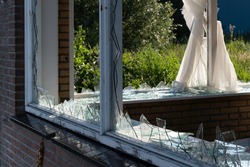 Broken glass windows, caused by vandalism or demolition, in a brick wall of a house. The curtains are partly down. Sharp points of glass stick from the rebate