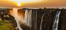 Wide panorama of victoria falls at sunset with orange sun in the sky and tourist in viewpoint