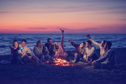 a group of friends using cell phones around the bonfire during autumn beach party drinking beer and having fun