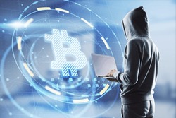 Hacker in hoodie using laptop computer with glowing bitcoin interface in blurry office interior. Hacking, theft and cryptocurrency concept. Double exposure