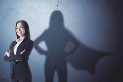 Attractive young european businesswoman with folded arms standing on concrete wall background with superhero cape shadow. Success and leadership concept