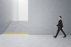 Young businessman walking in creative light concrete tile interior with stairs and mock up place on wall. School hallway and corridor concept