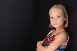 Little blonde girl with a serious expression on her face while standing against a black wall.