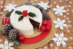 Christmas food still life with iced fruit cake, holly, snow covered winter greenery, fly agaric decorative mushroom  and white snowflake and red bauble decorations over oak background.