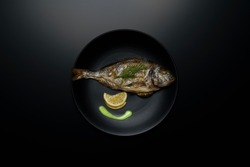 Grilled sea fish Dorada on a black plate and a black table, photo above