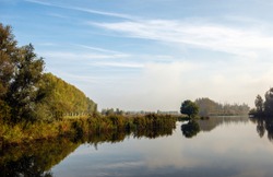 Trees and bushes around a Dutch lake with a mirror-smooth water surface. The photo was taken in autumn near the village of Lage Zwaluwe, North Brabant. Some haze is still visible in the background.