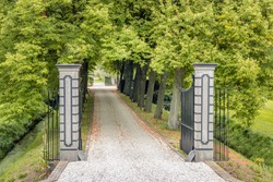 Opened black wrought iron gate between stone pillars. Behind the fence is a driveway covered with light colored gravel leading to an estate in the Netherlands. It is at the end of the summer season.