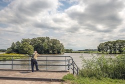 Unknown elderly man looks thoughtfully over the water of a Dutch lake while leaning on the bridge railing. He wears suspenders to hold up his blue jeans. It is a cloudy summer day.