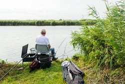 Unknown angler sitting on a chair for fishing in a small river. Around him are all kinds of accessories