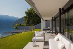 Private terrace overlooking Lake Ceresio in Switzerland. In front of a beautiful green lawn, small table, armchairs and sofa, all white. It's a sunny day and it's midsummer