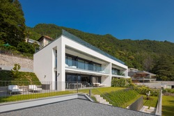 Modern house of two apartments with a beautiful garden directly on Lake Ceresio. Sunny day with blue sky. Minimalist and linear architecture. Panoramic view in background of green hills during summer.