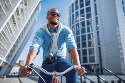 Low angle view of handsome young businessman in casual clothes and sun glasses riding his bike and smiling