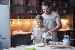 Cute little girl and her handsome dad in aprons are whisking eggs with flour and smiling while baking in kitchen at home