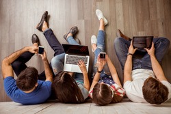 Group of attractive young people sitting on the floor using a laptop, Tablet PC, smart phones, headphones listening to music, smiling