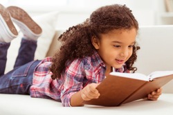 Cute little Afro-American girl in casual clothes reading a book and smiling while lying on a sofa in the room.