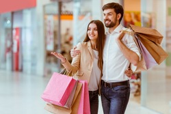 Happy beautiful young couple holding shopping bags, looking upon showcase and smiling while standing in mall