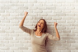 Happy beautiful young woman in casual clothes is keeping hands in fists and smiling, against white brick wall
