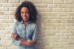 Beautiful Afro American girl is looking at camera and smiling while standing with crossed arms against white brick wall