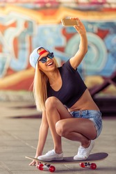 Beautiful blonde girl in glasses and cap is making selfie using a smartphone and smiling while standing on her skateboard in skate park